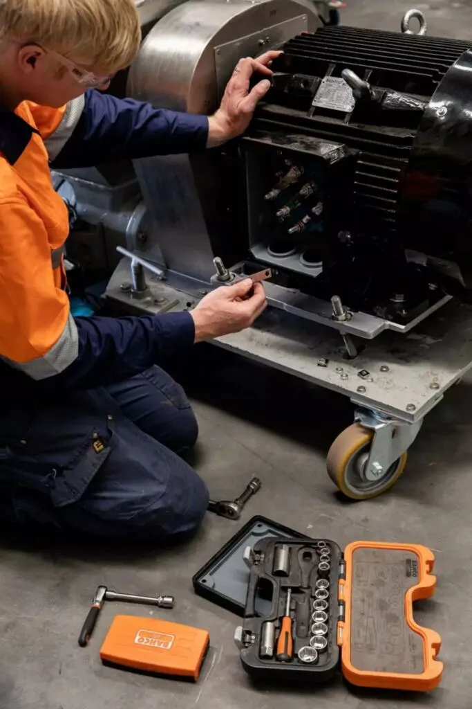 Technik w odzieży ostrzegawczej klęczy przy silniku elektrycznym, obok otwarta walizka z kluczami nasadowymi.