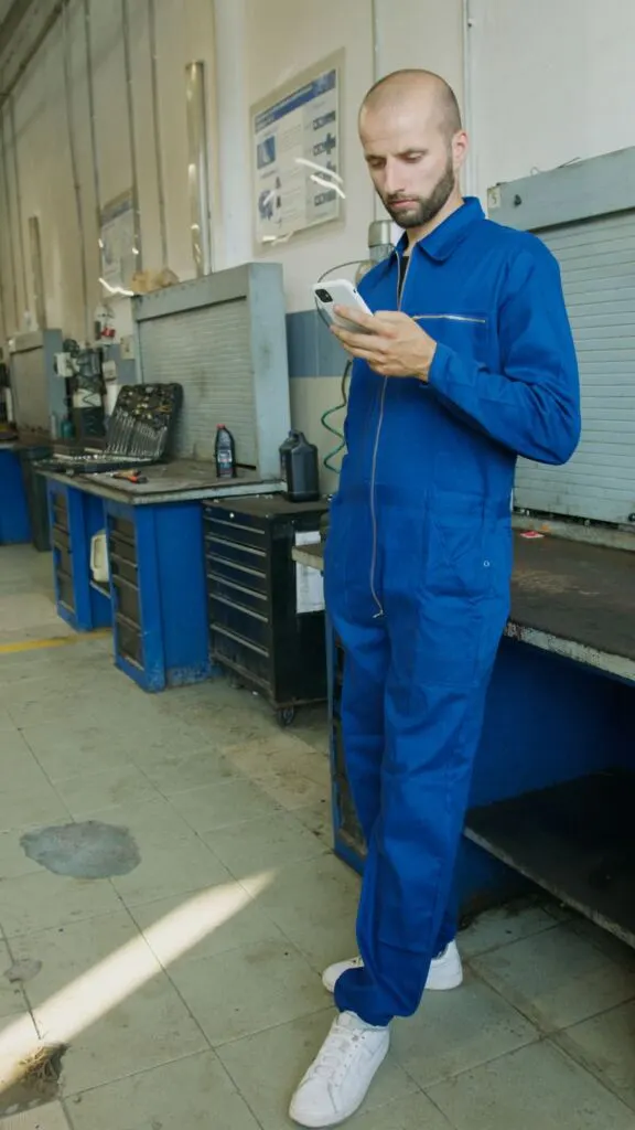 Mechanik w niebieskim kombinezonie z telefonem w ręku, w tle stoły warsztatowe z narzędziami.